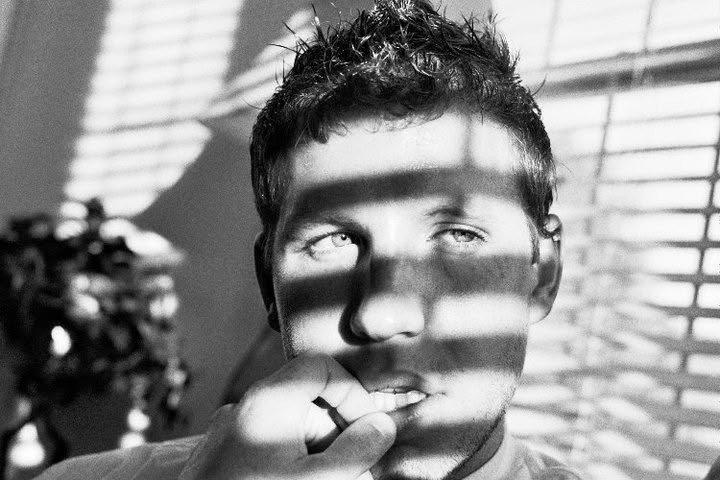 A young man’s face crossed by sharp bands of window-blind light