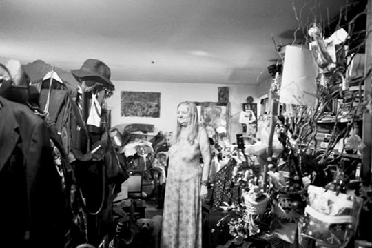A woman standing in a room crowded with clothing, objects, and branches