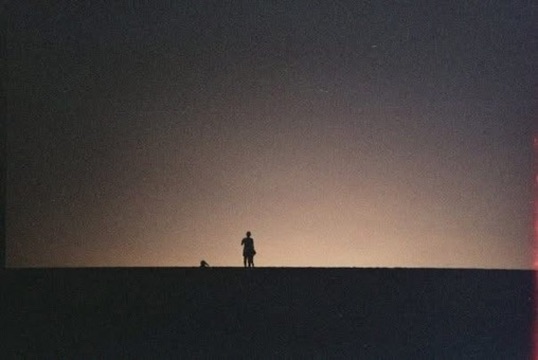 Two distant figures silhouetted along a dark horizon beneath a softly illuminated sky