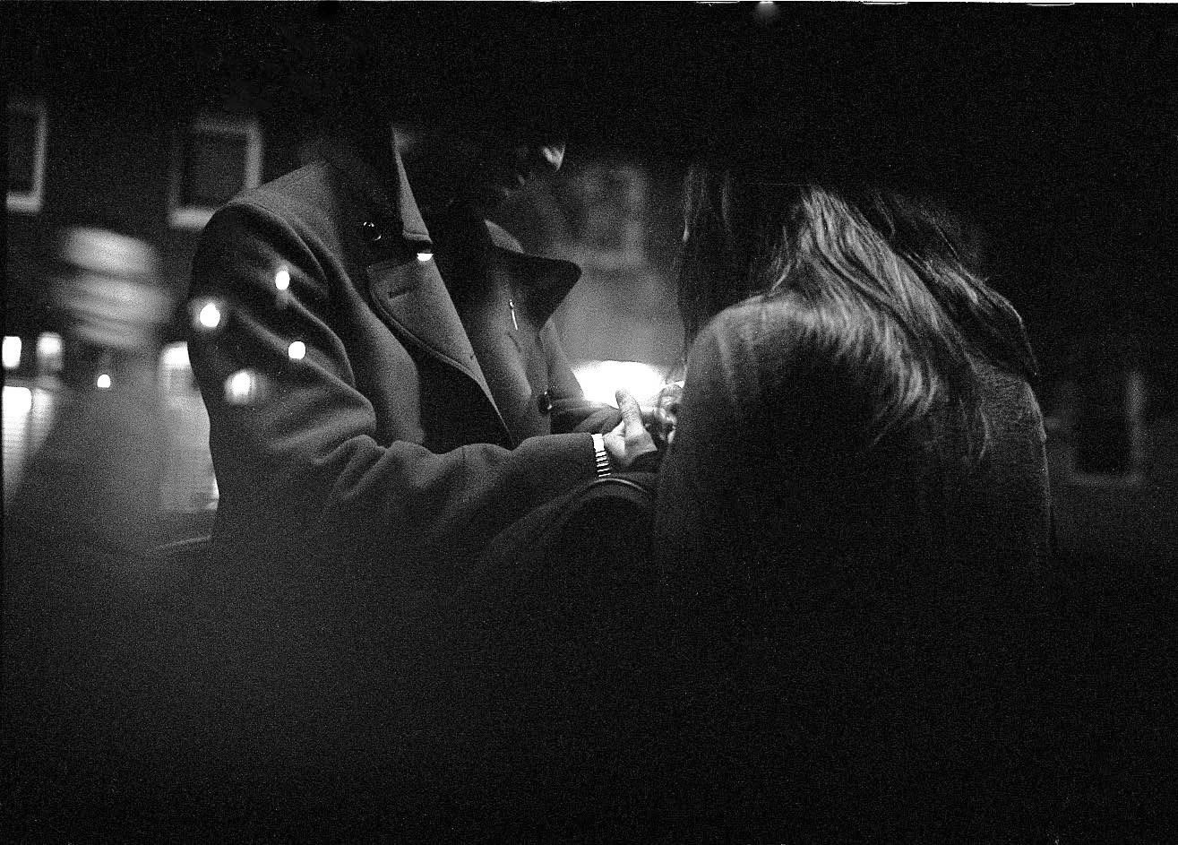 Two figures gathered closely on a city street at night, seen through reflections