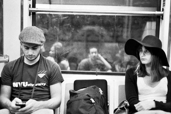 Two subway riders seated apart as a reflection appears in the window behind them