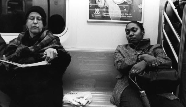 Two women seated apart on a subway bench, one reading and one listening through headphones