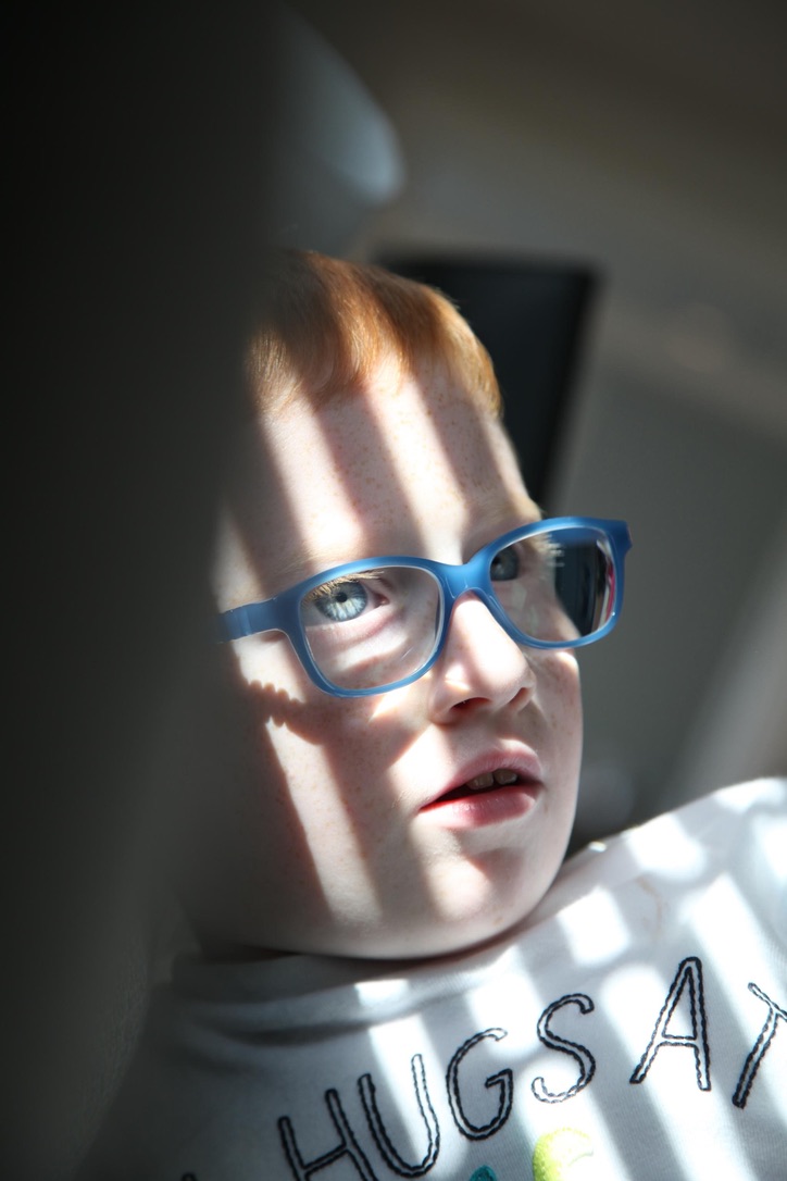 A red-haired boy in blue glasses crossed by bands of window light