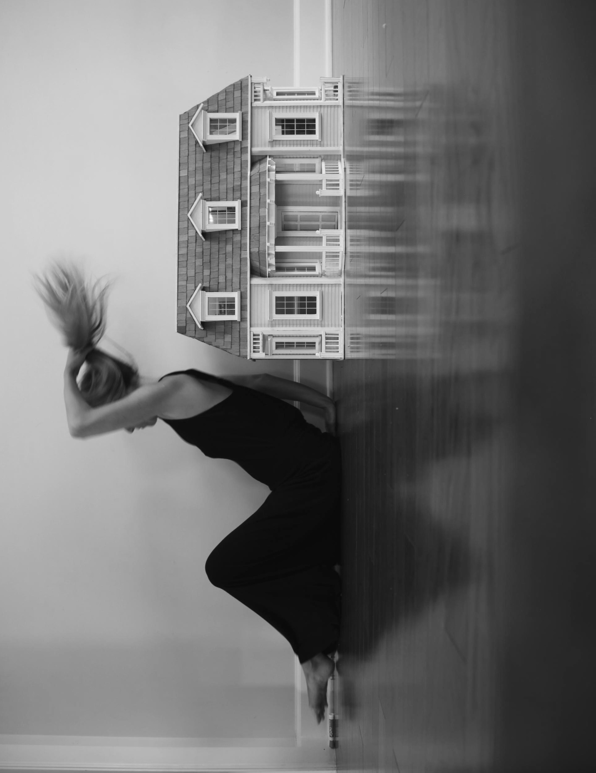 A woman and dollhouse arranged sideways within a tall black-and-white frame