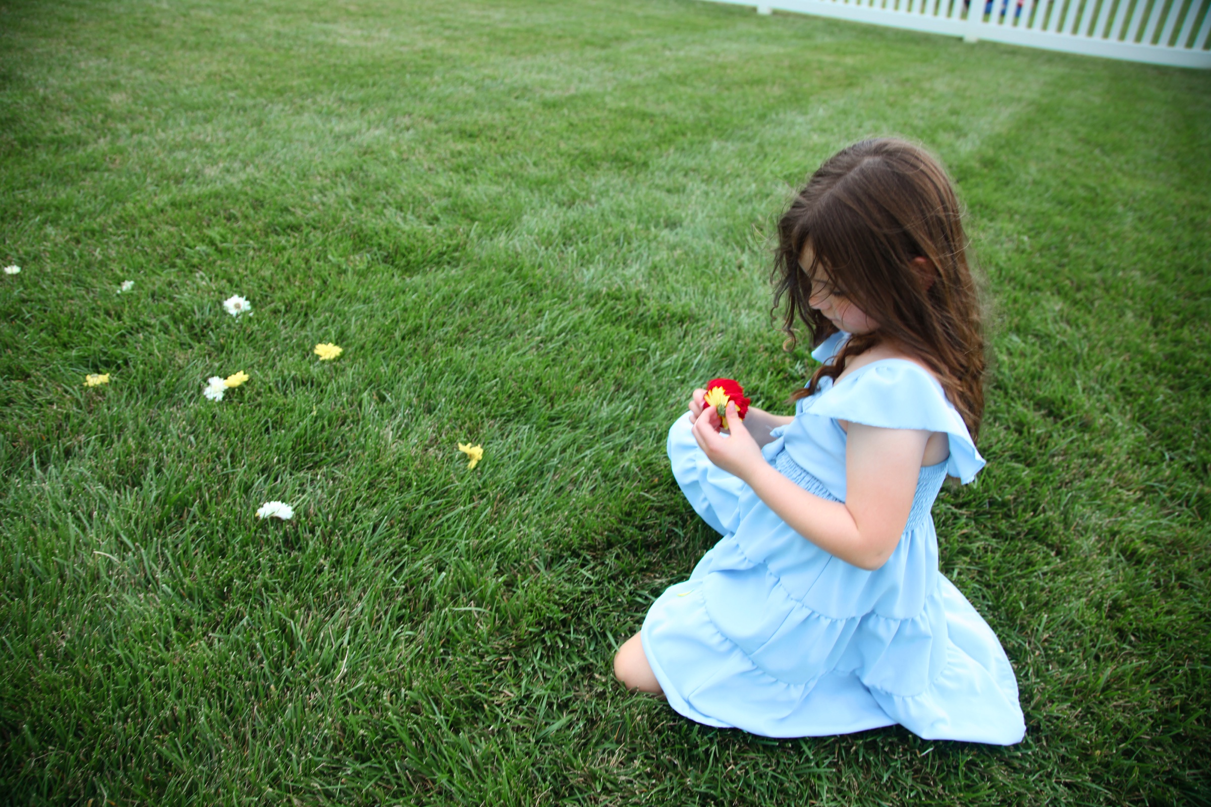 A girl in a pale blue dress kneeling in grass and holding a red flower