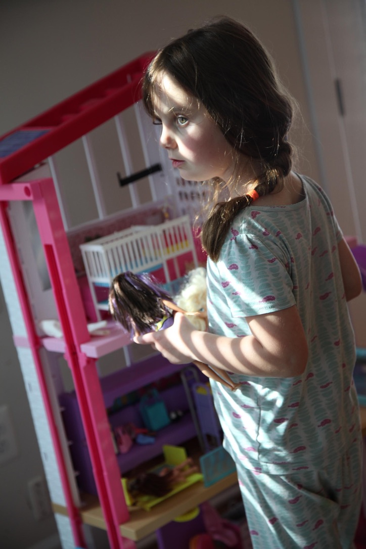 A girl holding dolls beside a pink dollhouse in bands of window light