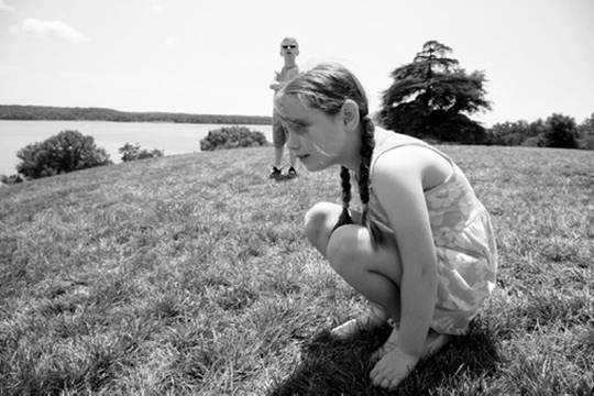 A girl crouching in grass near the water as an adult stands in the distance