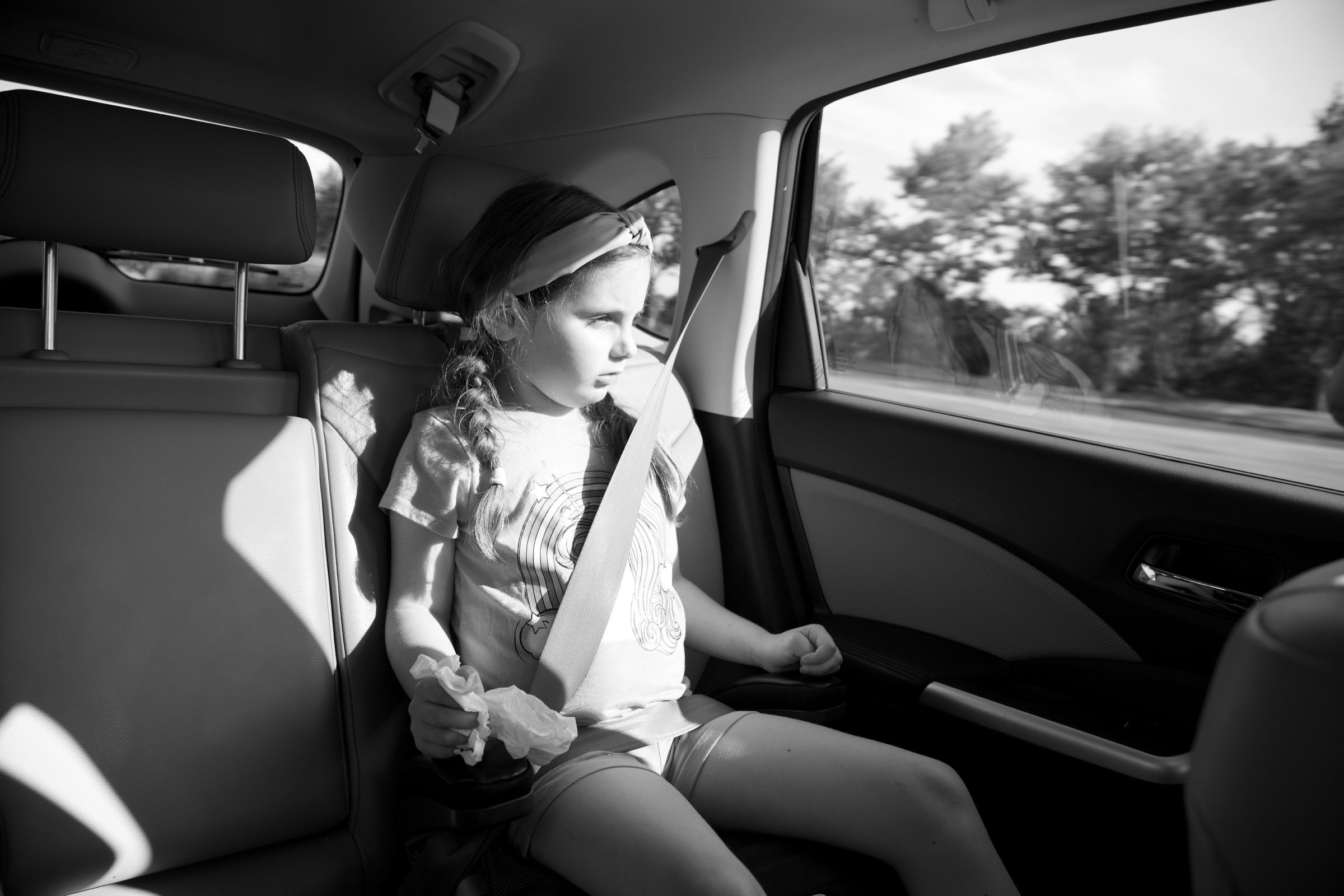 A young girl seated in the back of a car, lit by passing sunlight, in black and white