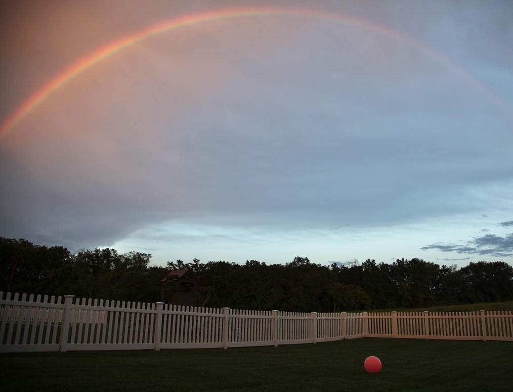 A rainbow arching over a dark yard, white fence, and solitary red ball