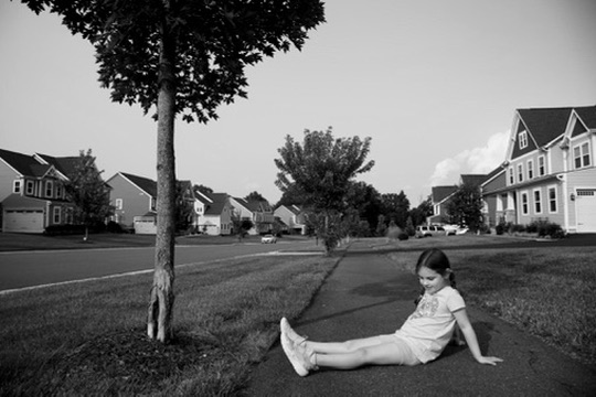 A girl seated alone on a sidewalk in a suburban neighborhood