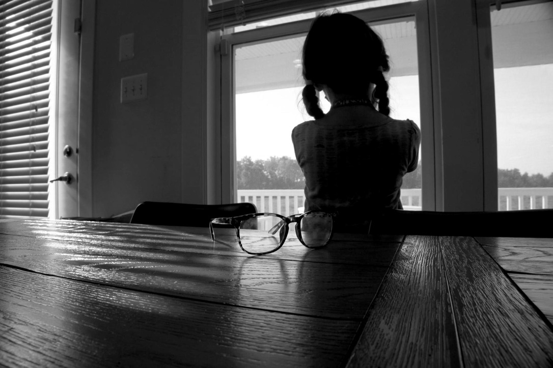 Eyeglasses on a wooden table before a child silhouetted at a bright window